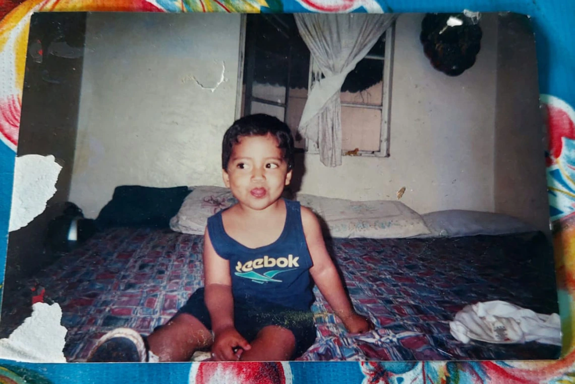 Old photo of a young child with dark hair sitting on a bed, goofily looking sideways and sticking out his tongue