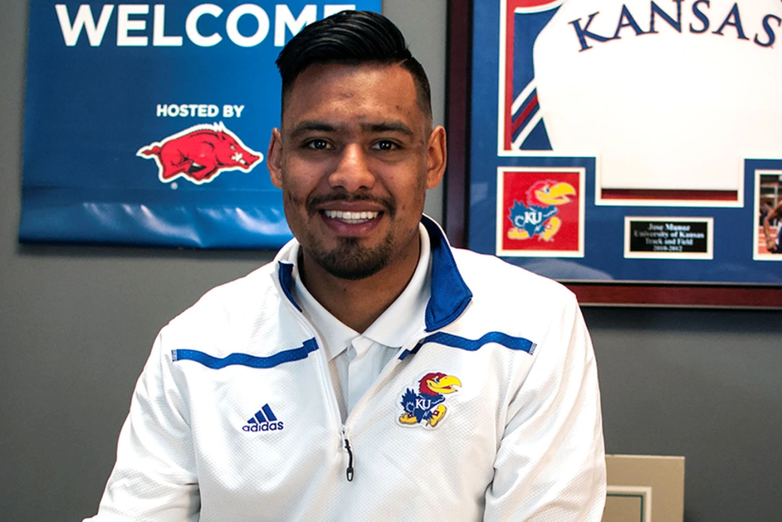 Young man in a white long-sleeved shirt sits smiling among University of Kansas memorabilia 