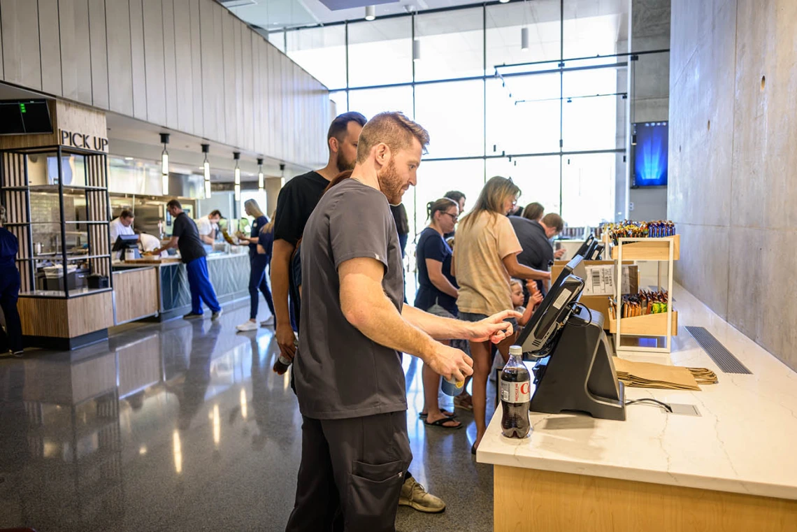 Several people stand at computer screens in a cafe setting placing their orders. 