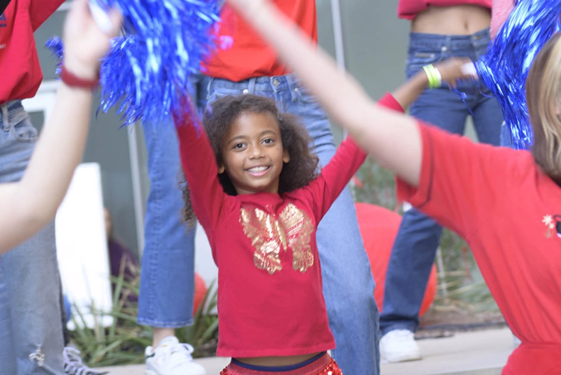 A group of people of mixed ages all wearing red and blue are smiling and dancing waiving pom poms.