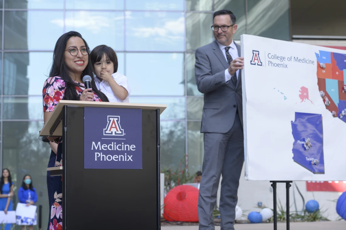 A young adult woman with long dark hair stands at a UArizona podium holding a child. 