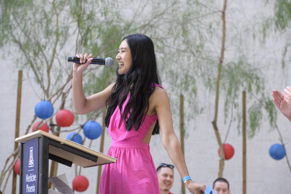 A young Asian woman with long dark hair speaks into a microphone in an outdoor setting. 