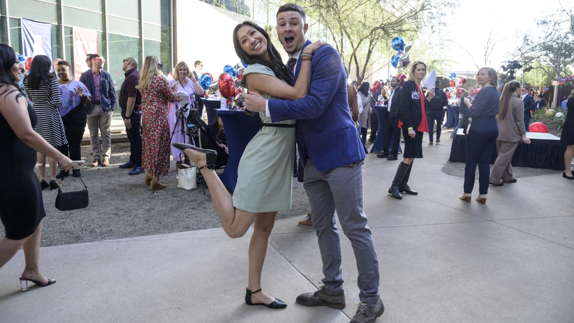 Young adult man and woman embrace, looking at viewer smiling and woman has her leg bent up behind her. 