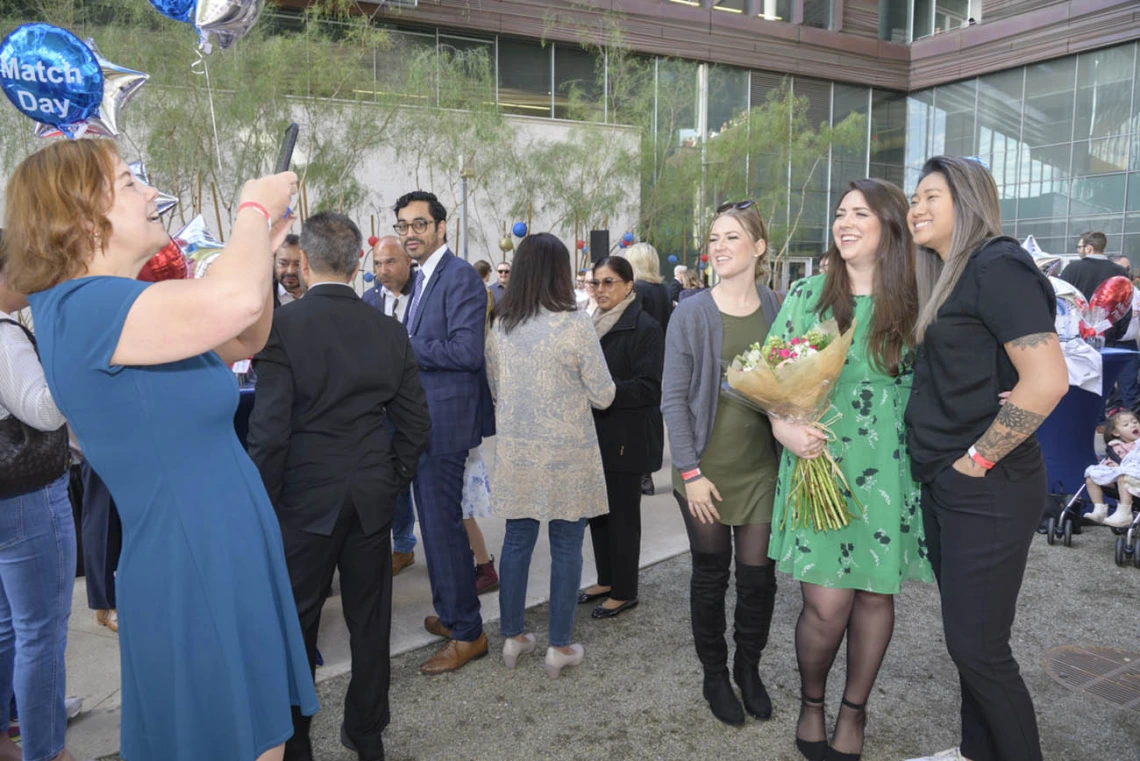 A woman with red hair takes a photo of three woman standing next to each other smiling. The woman in the middle is holder flowers. 