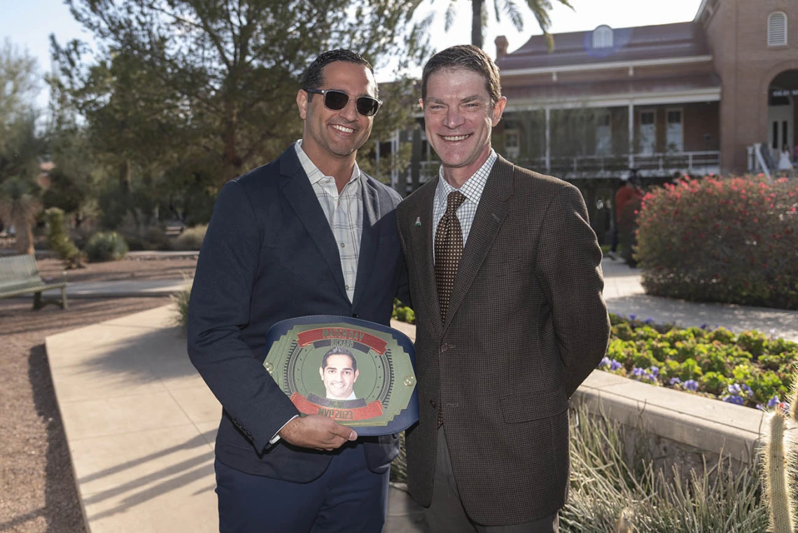 Two men in suites stand outside smiling. The younger man with dark hair holds an oval plackard with his photo on it. 