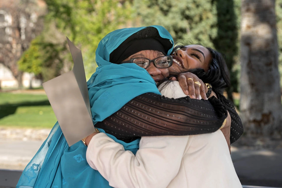 A young woman is hugged by her mother as they both smile. Both have dark skin and the young woman is holding a letter. 