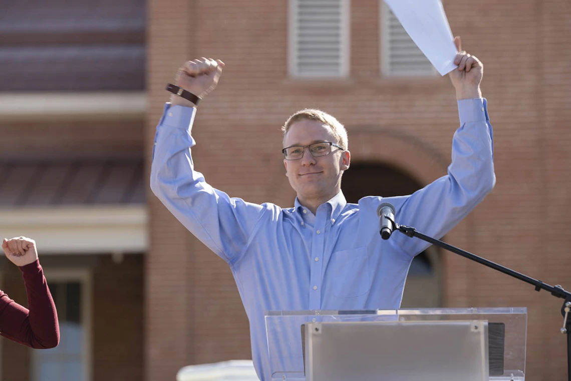 A smiling young adult man with light skin and hair in a dress shirt holds up both arms in celebration. 