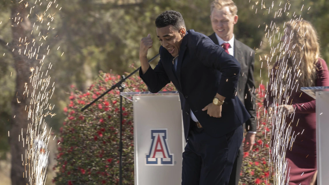 A young adult male wearing a dark suite does a dance on a stage with sparklers going off around him. A University of Arizona logo is behind him. 