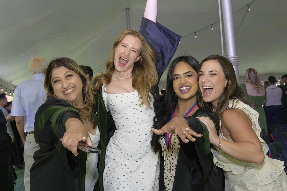 Four young women in graduation regalia smile and hug in celebration. 