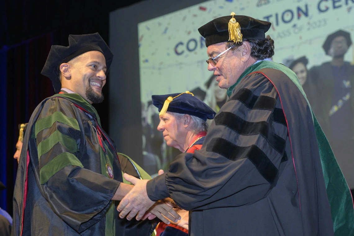 A smiling young man in graduation regalia shakes hands with the dean of the college. 