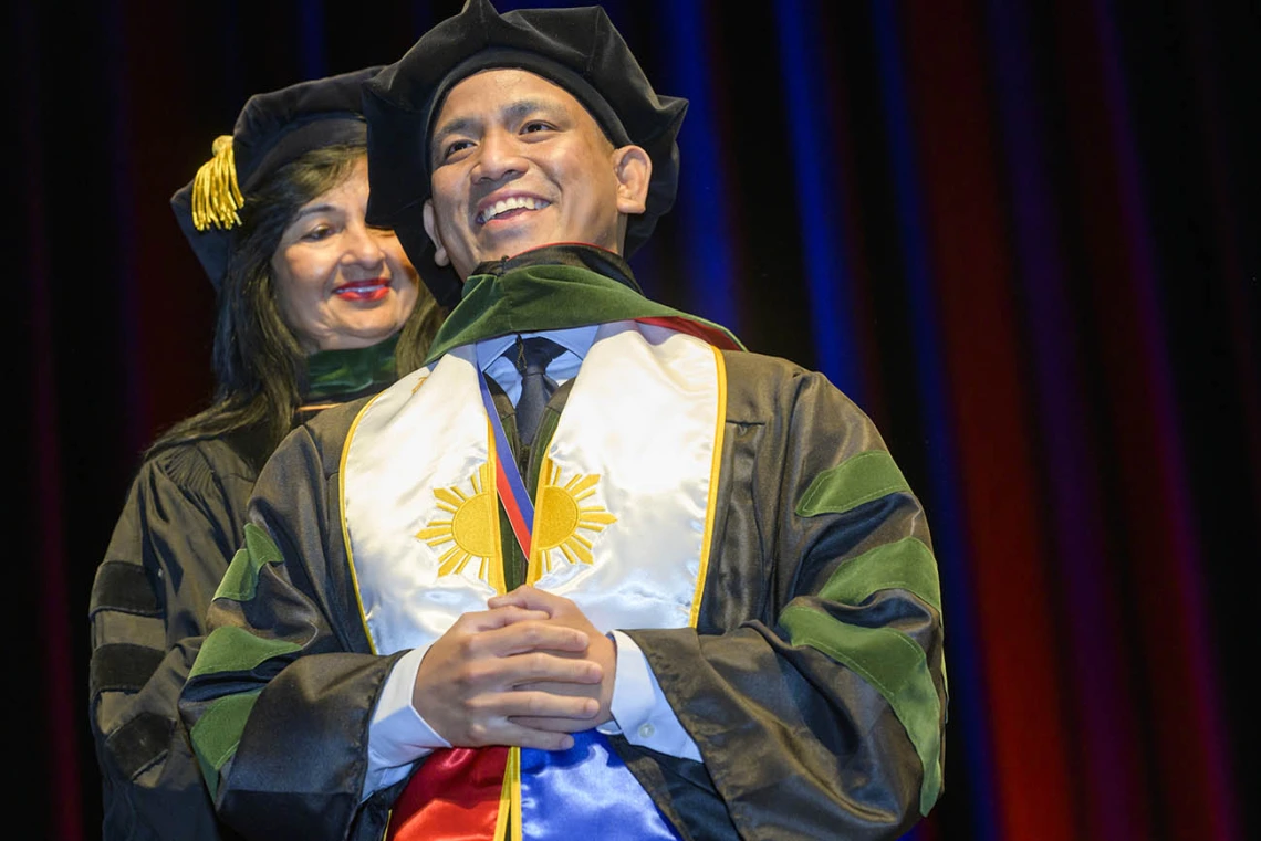 A young man in graduation regalia with a white stohl around his neck, smiles as a professor places a sash over his shoulders. 