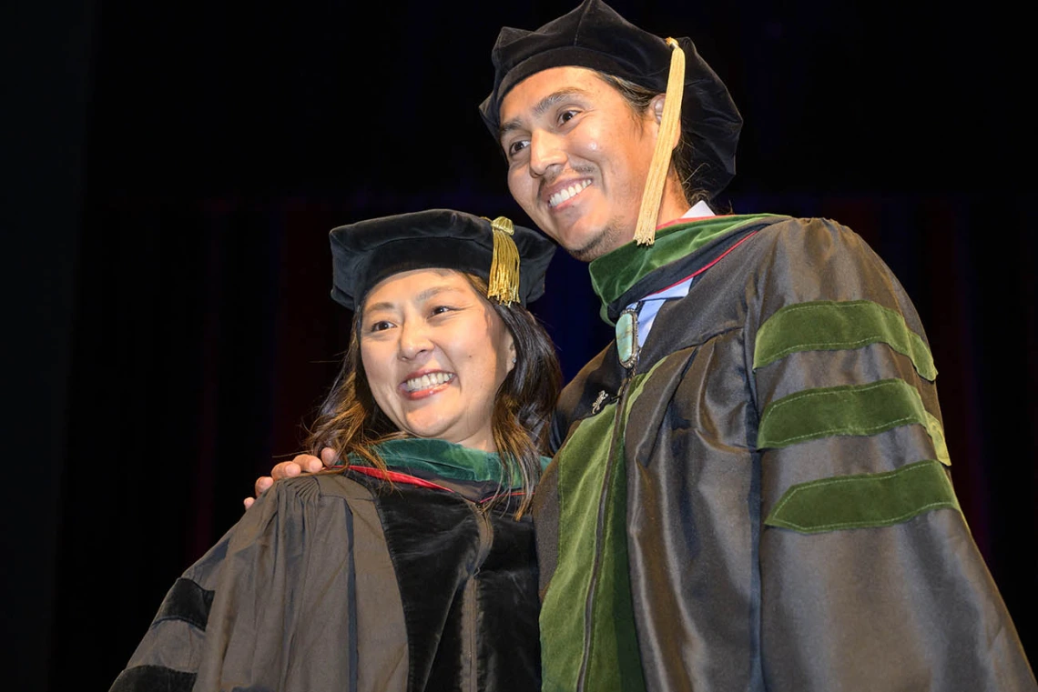 A tall young man in graduation regalia side hugs a professor, also in graduation regalia, as both smile. 