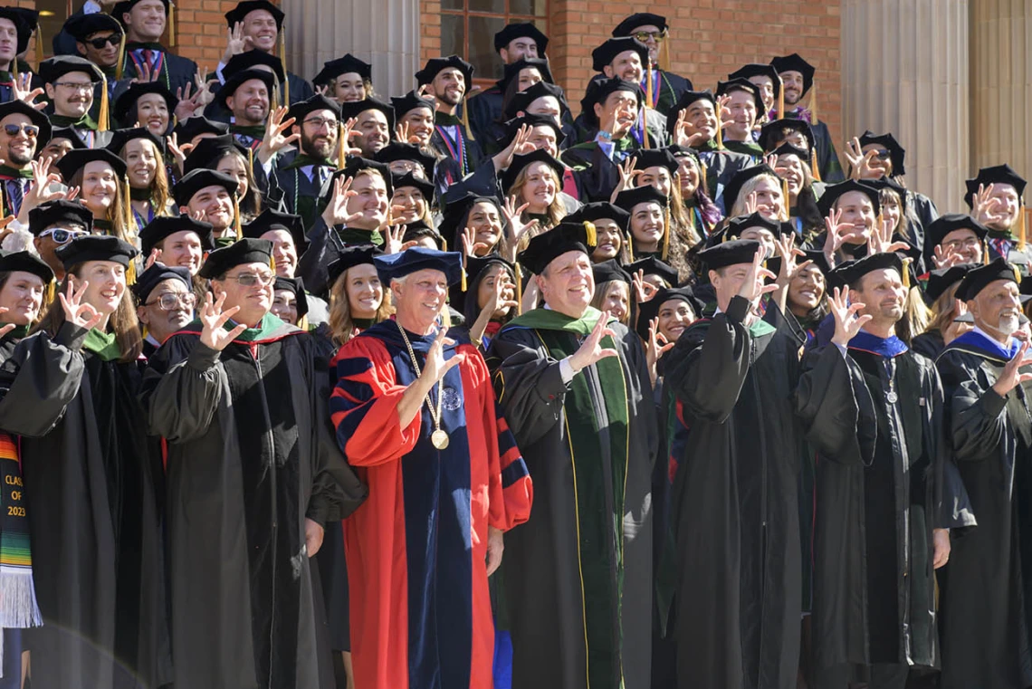 Dozens of graduates and faculty in graduation regalia stand on stairs making the Arizona Wildcat sing with their fingers. 