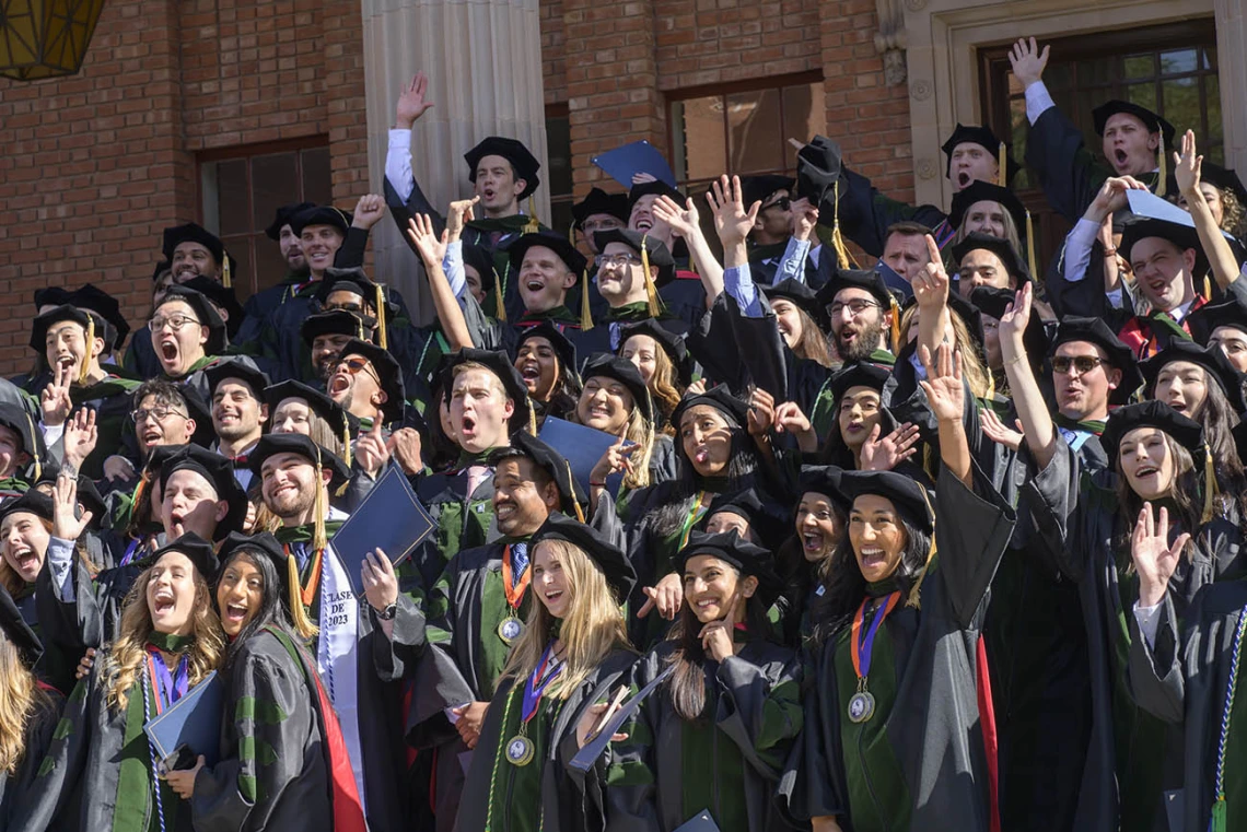 Large group of medical students in graduation robes and hats wave and cheer. 