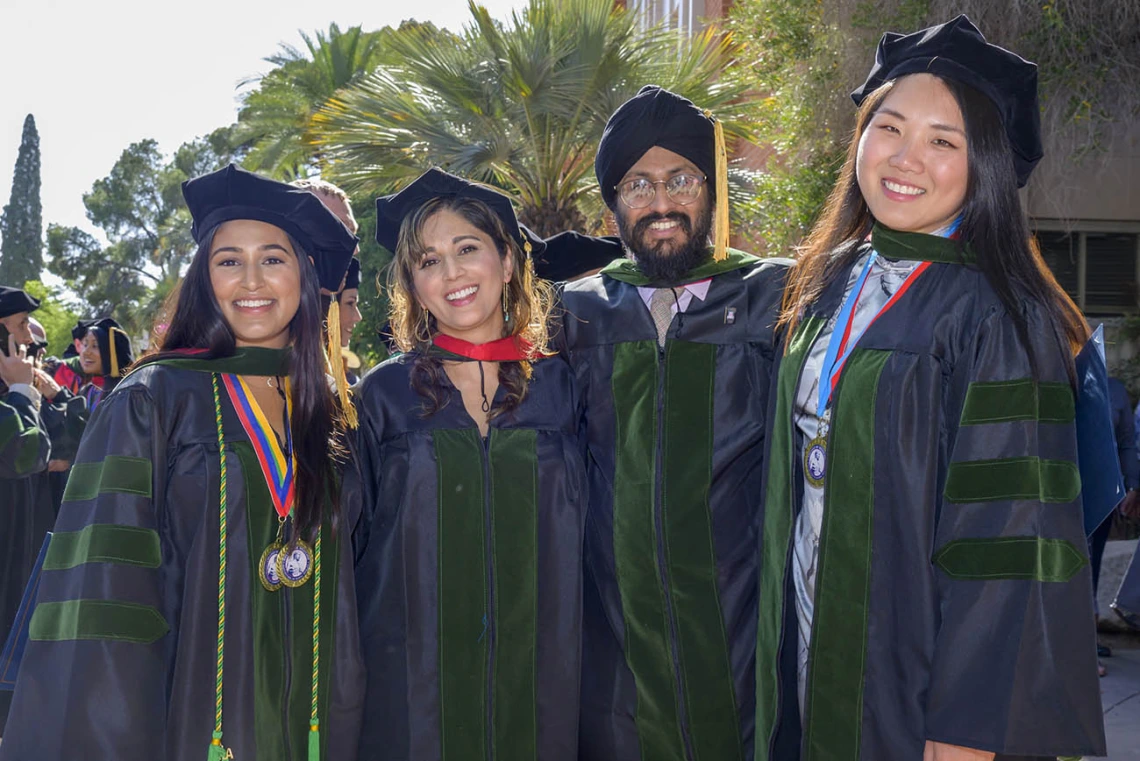 Four medical school graduates in caps and gowns stand together smiling. 