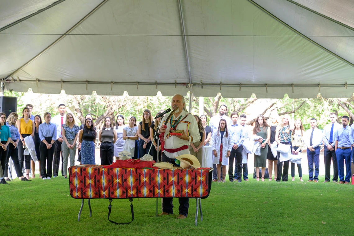 An oudoor photo of a Native American man standing at a table leading a blessing ceremony with a croud of people standing around him. 