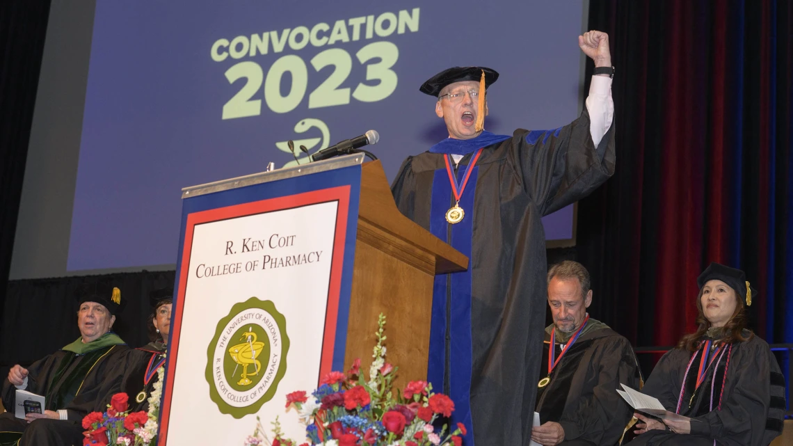 Older white man dressed in graduation cap and gown stands at a podium witih fist in air as if cheering. 