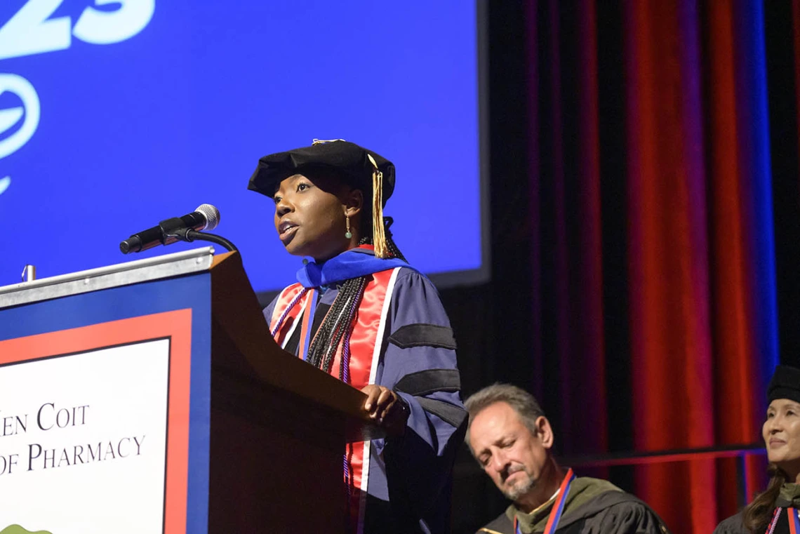 A young Black woman in graduation cap and gown talks into a microphone at a podium. 