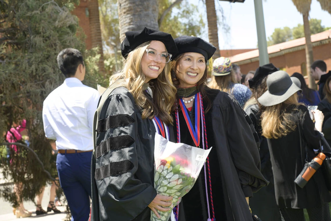 A young woman with long blonde hair in a graduation cap and gown smiles, holding flowers and posing with a female professor in graduation regalia as well. 