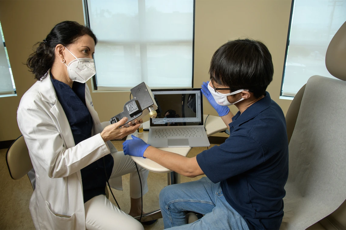 Clara Curiel Lewandrowski, MD, talks with Dongkyun Kang, PhD, as she holds the portable confocal microscope the two are developing to provide advanced skin cancer diagnostic options in the clinic. The portable device will help doctors diagnose and excise skin cancer while reducing the wait for biopsy results.