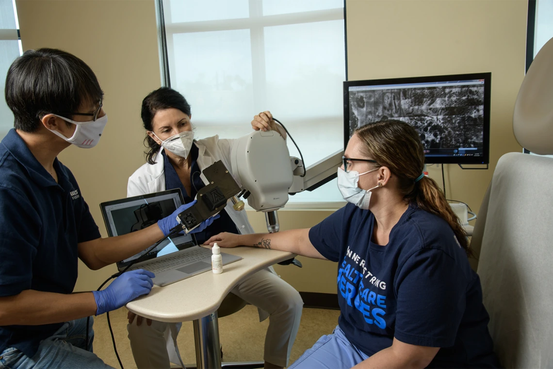 Clara Curiel Lewandrowski, MD, and Dongkyun Kang, PhD, prepare to do a side-by-side test of the confocal microscope and the portable unit to compare the image quality. Dr. Curiel is aligning the larger device, while Dr. Kang prepares to aim the handheld device at a nearby spot on Savannah Barber’s arm. Barber is a medical assistant at Banner – University Medical Center.