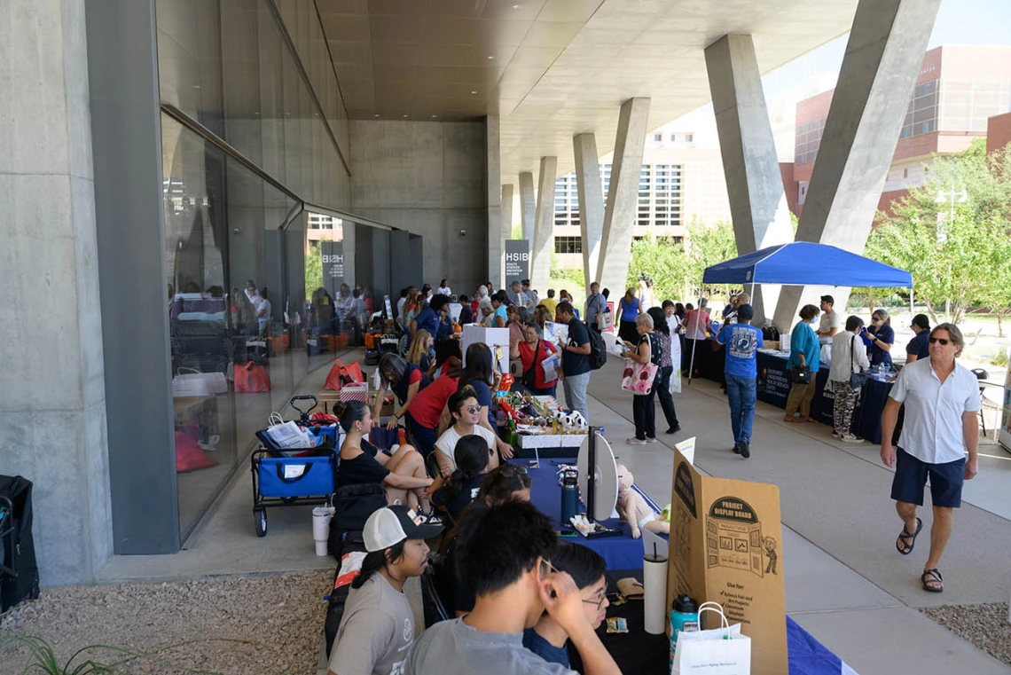 A crowd of people mill about information tables in an outside patio next to a building. 