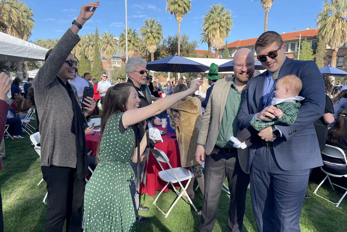 A light-skinned man in a suite holds a baby while several people are trying to get the baby to look their way for a photo.