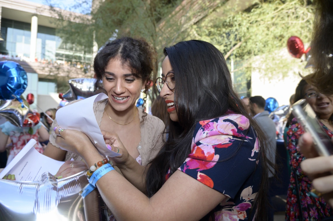 Two young adult women with dark hair look at each other's letters and smile. 