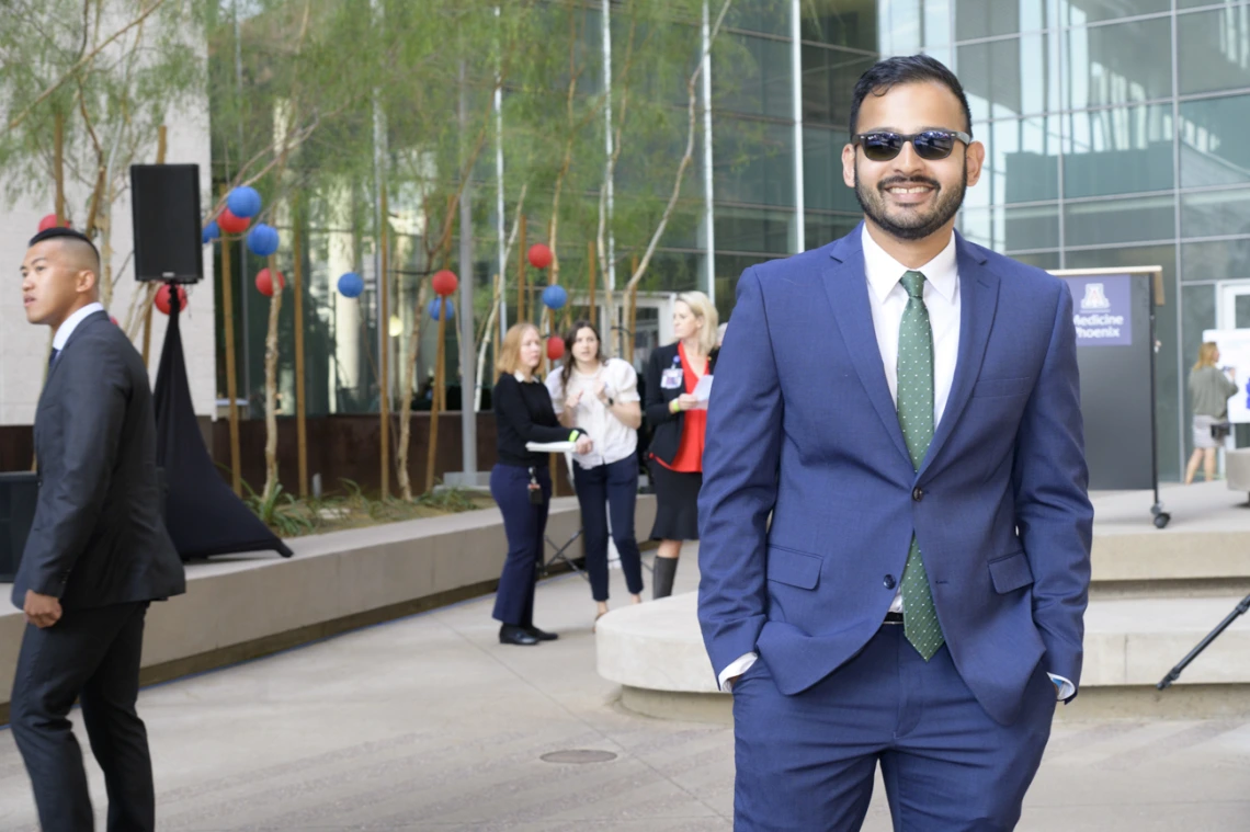 A young adult man in a suite and tie with short dark hair, sun glasses and a beard smiles in an outdoor setting. 