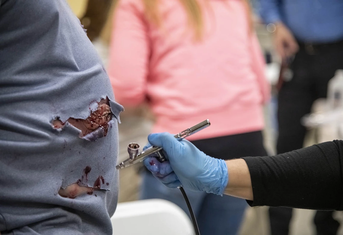 Deana Ann Smith, BS, BSN, RN, healthcare simulation educator for ASTEC, air brushes blood-red special-effects makeup on a man’s stomach for an emergency exercise. 