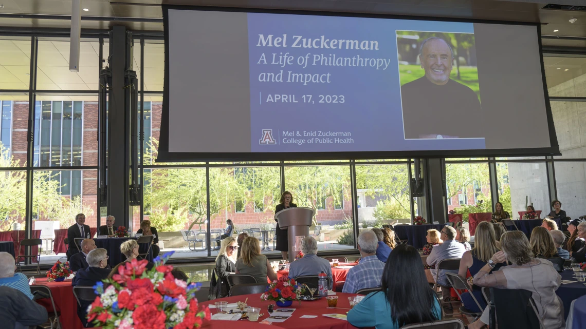 Crowd of people sitting at tables watching Dr. Iman Hakim, dean of the University of Arizon Mel and Enid Zuckerman College of Public Health, speak at a podium.