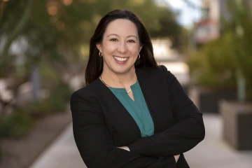 Portrait of woman with dark brown hair smiling with her arms folded in front of her. She is wearing a teal blouse and a black blazer.