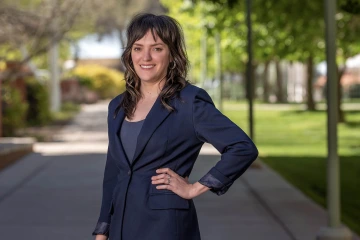 Woman with brown hair and bangs waring a navy blue blazer stands outside smiling with one hand on her hip.