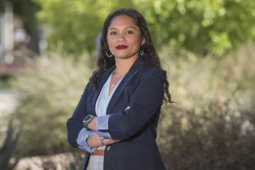 Woman with long, dark, curly hair wearing a blue blazer and shirt standing outside.