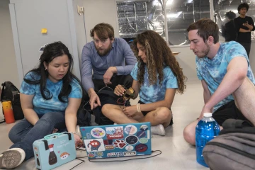 Three students in tie-dyed shirts sit on the floor looking at a laptop while holding a bioengineered device with their professor kneeling down talking with them.