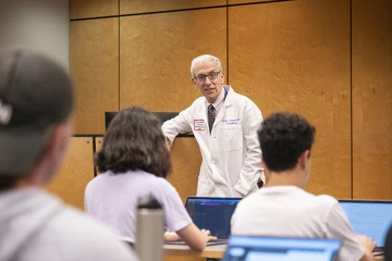 Older white male wearing glasses and a white coat stands at the front of a class giving a lecture to students.