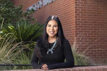 A smiling woman with long brown hair, wearing a black long sleeve top and turquoise necklace, stands in front of a red brick building.