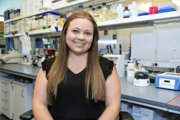 Woman with long brown hair wearing a black blouse smiling in a lab.