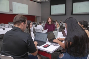 Bridget Murphy visits with students at a table in a large classroom.