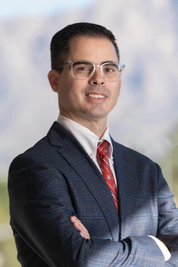 Portrait of an Hispanic man with short brown hair wearing glasses, a dark suit and a red tie. He is standing with his arms crossed and there is a mountain range behind him.