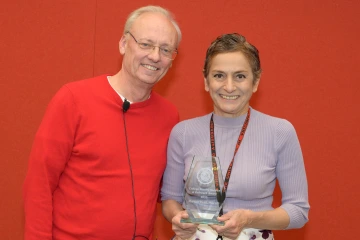 A woman and man stand next to each other holding a glass award between them, both smiling.