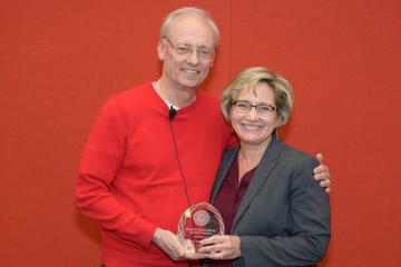 A woman and man stand next to each other holding a glass award between them, both smiling.