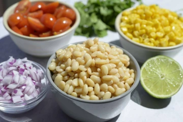 A kitchen counter with bowls of tepary beans, red onions, corn, and cherry tomatoes, with half a lime and cilantro ready to make a bean salad