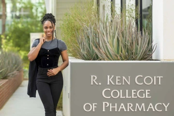 Outside portrait of Brena Andrews standing alongside the sign for the University of Arizona’s R. Ken Coit College of Pharmacy. 