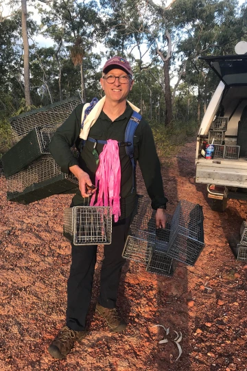 A middle-aged white man stands outside a truck holding several small metal cages.