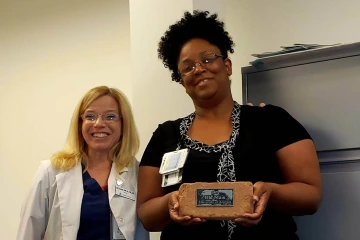 Taller Black woman, Jocelyn Nelms, wearing a black blouse and holding a box stands beside a shorter white woman, Deborah Gorombei wearing a white coat with her hand on Jocelyn's shoulder.