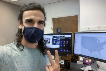 A man with shoulder-length brown hair and holding up a peace symbol with his fingers, stands in front of a dual-screen computer monitor. 