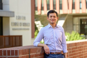 Portrait of an Asian man wearing a dress shirt standing outside leaning against a half-brick wall 