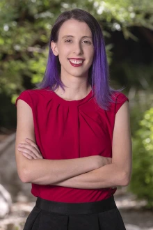 Young woman with brown and purple hair stands smiling with her arms folded. She is wearing a white short-sleeved blouse.