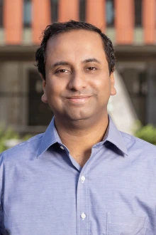 Portrait of an Indian man with short dark hair wearing a blue dress shirt in an outdoor setting. 